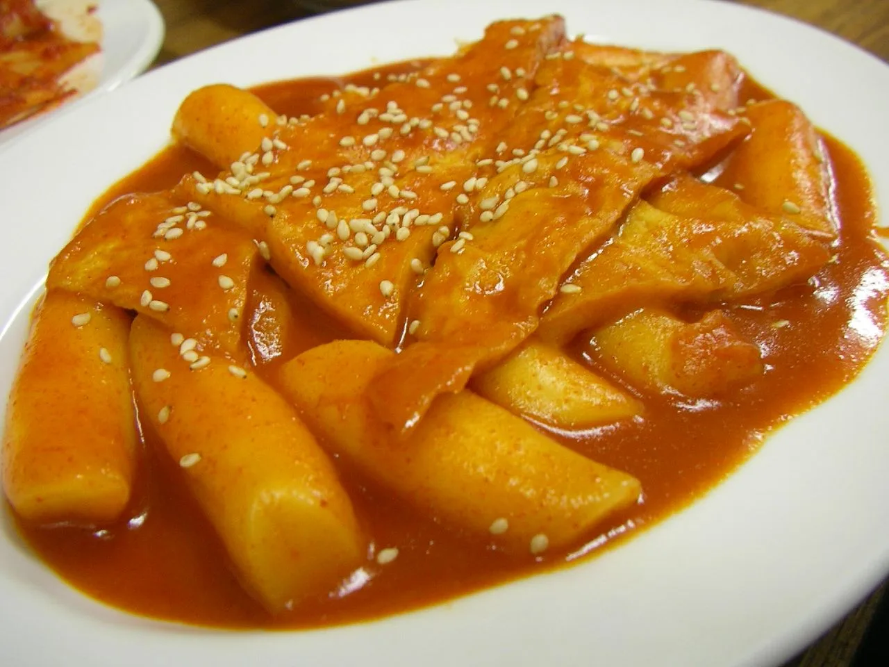 The post-war red version: rice cakes and fish cake in gochujang sauce, the style Sindang-dong made standard. The recipe below is the older soy-sauce one. Tteokbokki on a white plate — rice cake sticks and sheets of fish cake in red gochujang sauce, topped with sesame seeds