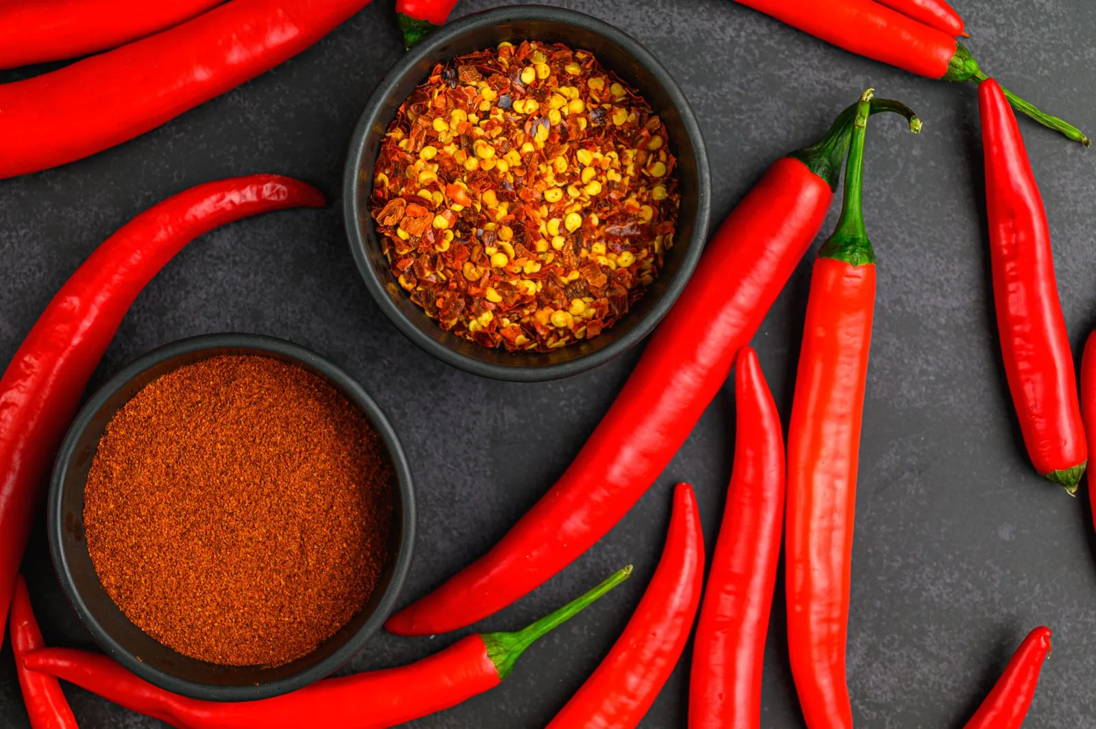 Coarse chili flakes and fine chili powder in dark bowls with fresh red chillies