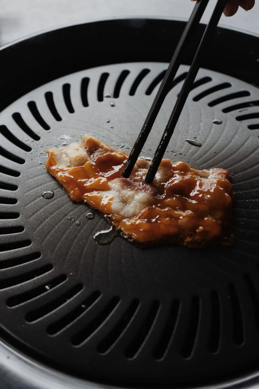 Close-up of marinated pork slice being grilled on a Korean barbecue grill using chopsticks.