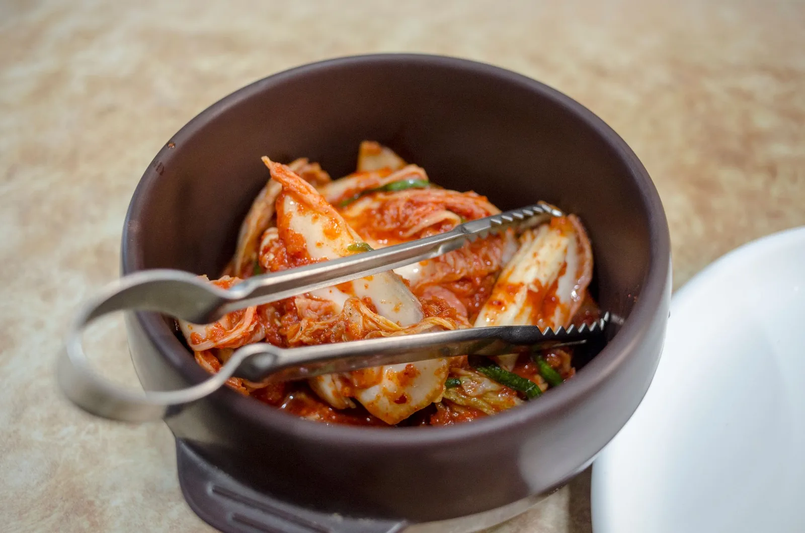 Close-up of traditional Korean kimchi in a dark ceramic bowl with tongs.