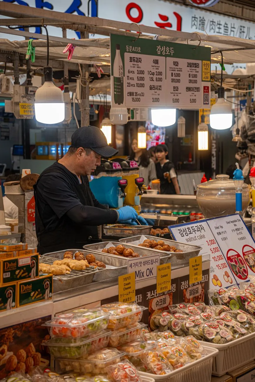 A vibrant market scene with a street food vendor preparing Korean dishes under bright lights.