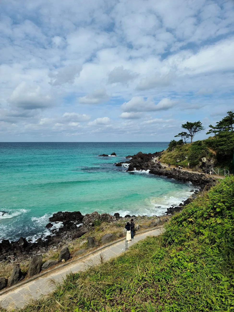 A picturesque view of a turquoise sea and rocky coastline in Jeju, South Korea, under a partly cloudy sky.